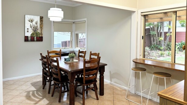 A dining area with a wooden table and chairs, a hanging light fixture, a clock on the wall, and a window with a view of a garden. - Buller Street Home (Charlestown)