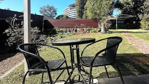 An outdoor patio with a table and two chairs, a covered area, and a view of a building and trees in the background. - Buller Street Home (Charlestown)