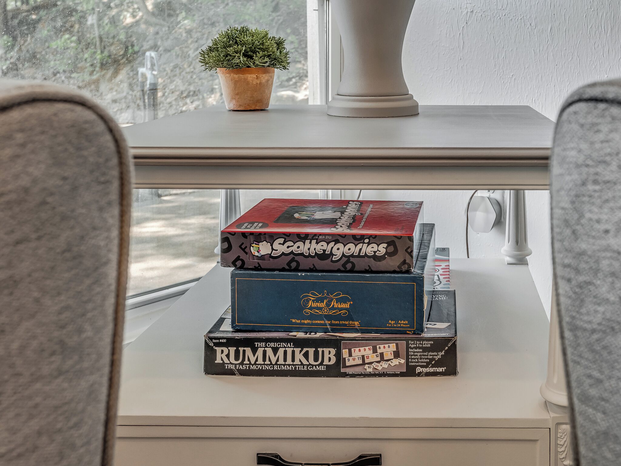 Une pile de jeux de société est posée sur une table blanche, près d’une fenêtre.
