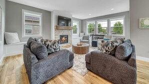 A spacious living room with a fireplace, two grey sofas with patterned cushions, a round coffee table, and large windows offering a view of greenery outside. - Trailside Treasure | Pool | Near Lake and Downtown | Hiking & Biking Trail (South Haven)