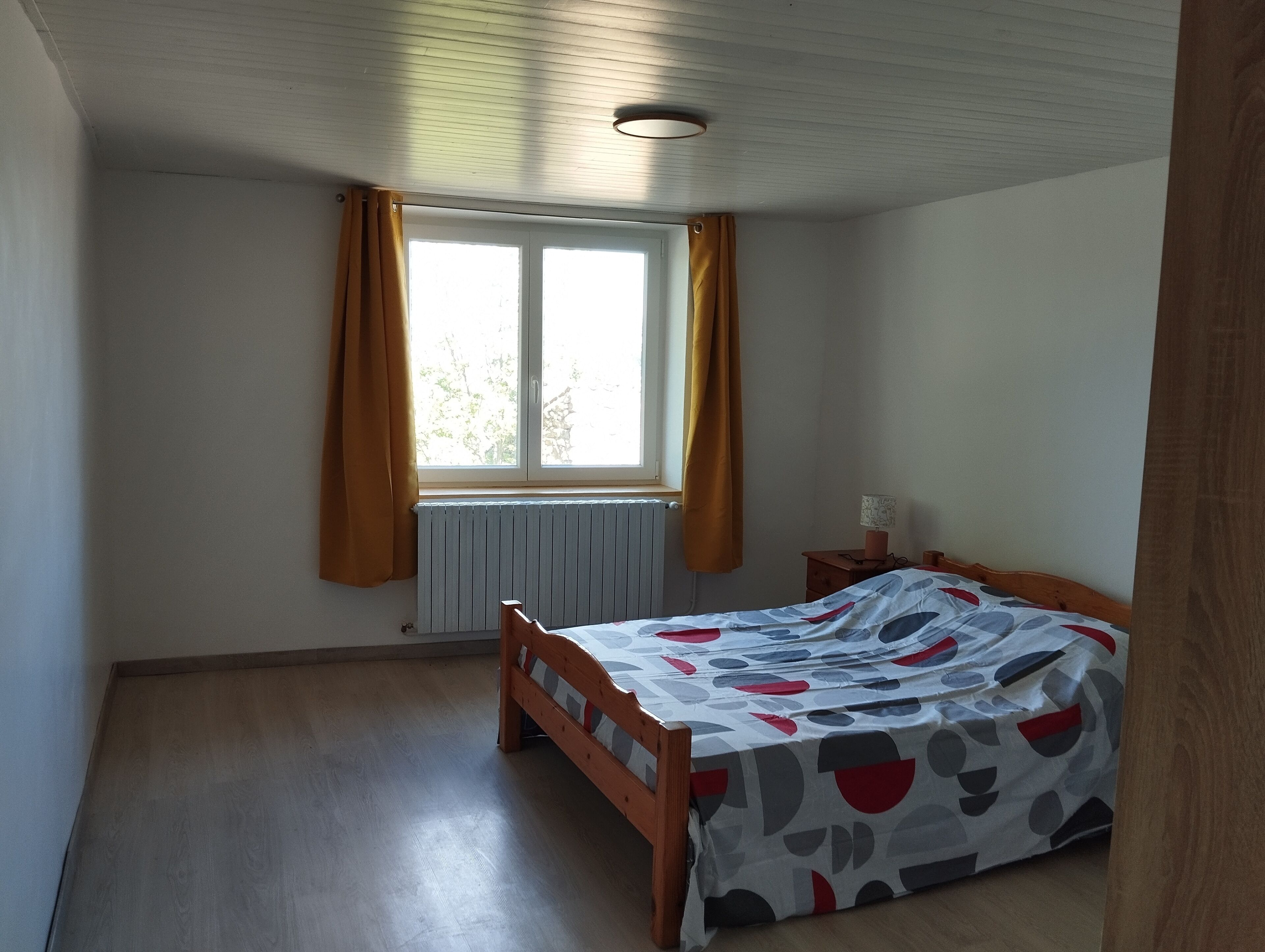 Una camera da letto con un letto in legno, una finestra con tende, un termosifone e un comodino con una lampada.