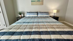 A neatly made bed with a striped bedspread, a small bedside table with a lamp, a framed picture on the wall, and a potted plant. - Villa Escondida #2 -2BD- Straight Shot to SpaceX! (Brownsville)