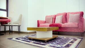 Una sala de estar con un sofá rosa, un sillón blanco, una mesa de centro de madera y una alfombra estampada.
