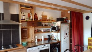 Une cuisine compacte équipée d’une cuisinière, d’un lave-vaisselle, d’un four et d’un lave-linge. On y trouve également un petit réfrigérateur, une tablette posée sur un support et divers ustensiles de cuisine rangés sur des étagères.
