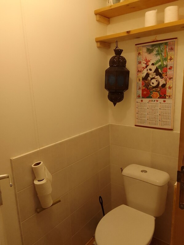Une petite salle de bain comprenant des toilettes, une étagère avec du papier toilette, un calendrier et une lanterne décorative.
