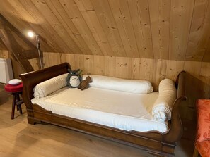 A wooden bed in a room with a sloped ceiling, a bedside table with a lamp, and a red stool.