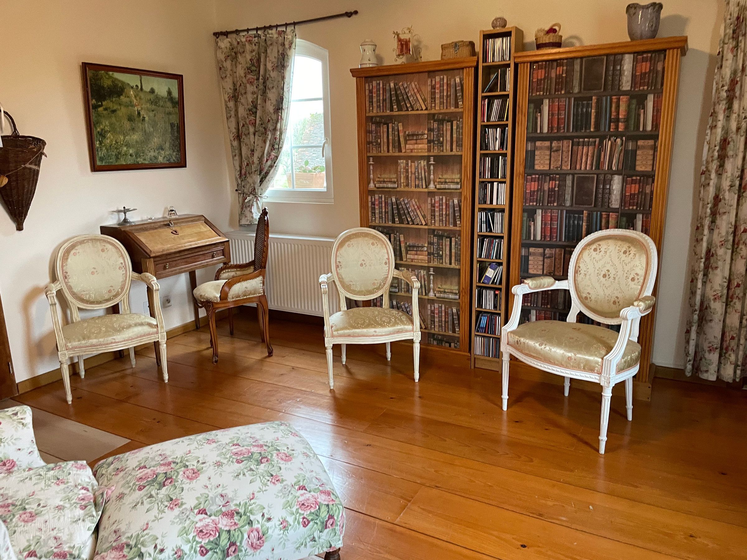 A room with wooden flooring, a bookshelf, two chairs, a small table, and a window with curtains.