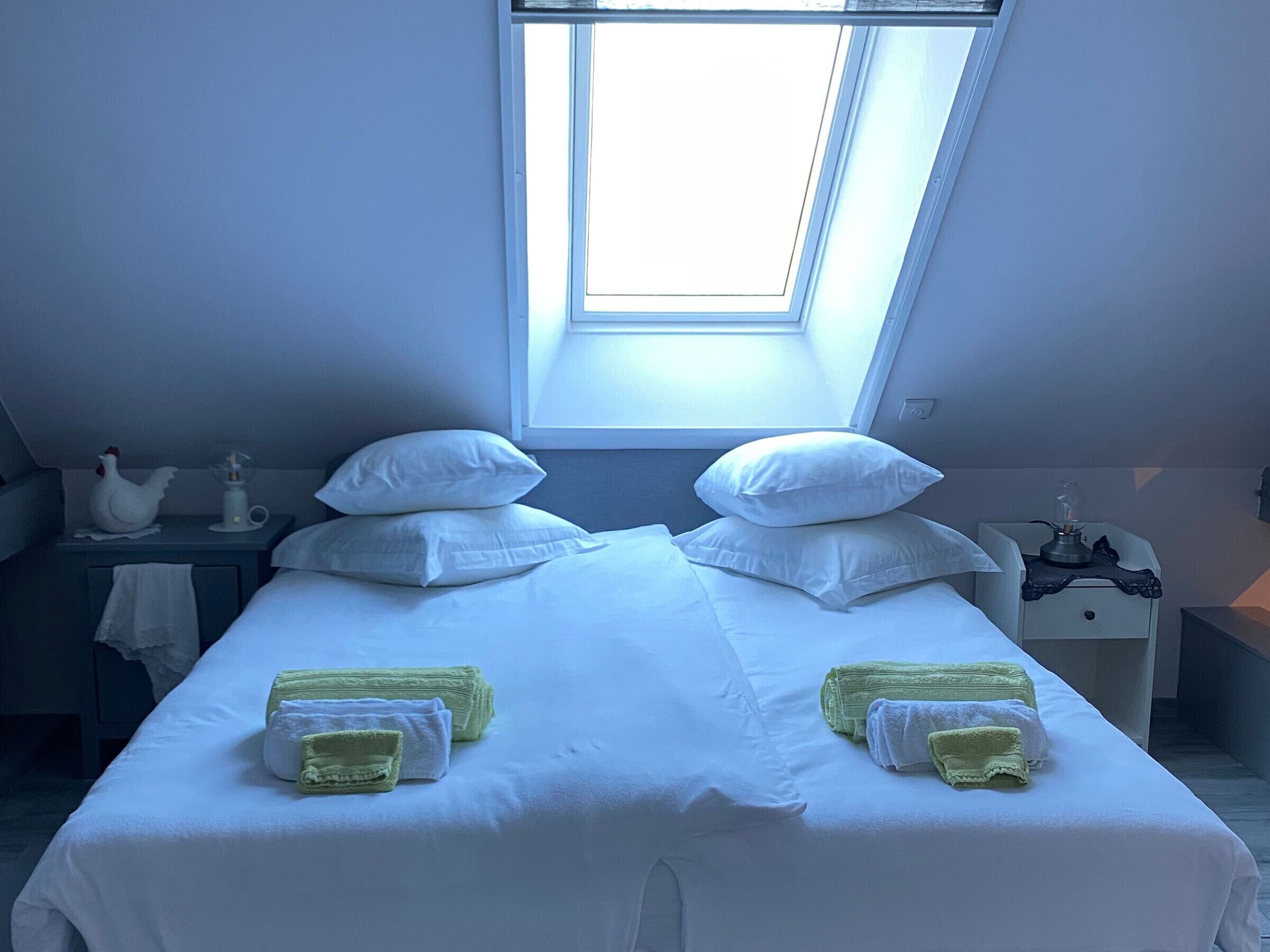 Una cama doble con ropa de cama y almohadas blancas, una mesita de noche pequeña con una lámpara, y una ventana sobre la cama.
