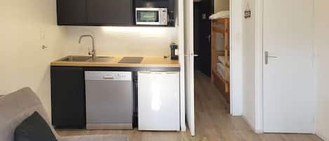 Une cuisine compacte équipée d’un lave-vaisselle, d’un four à micro-ondes et d’un réfrigérateur. On y trouve également un petit coin salon avec un canapé et une plante en pot.