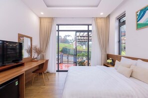 A hotel room with a large bed, a desk with a television, and a balcony with a view of the garden.