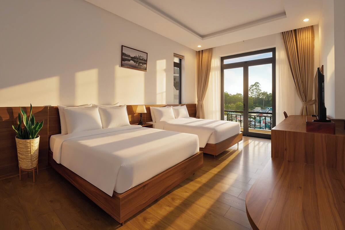 A hotel room with two beds, a wooden floor, a large window with curtains, a plant, and a TV.