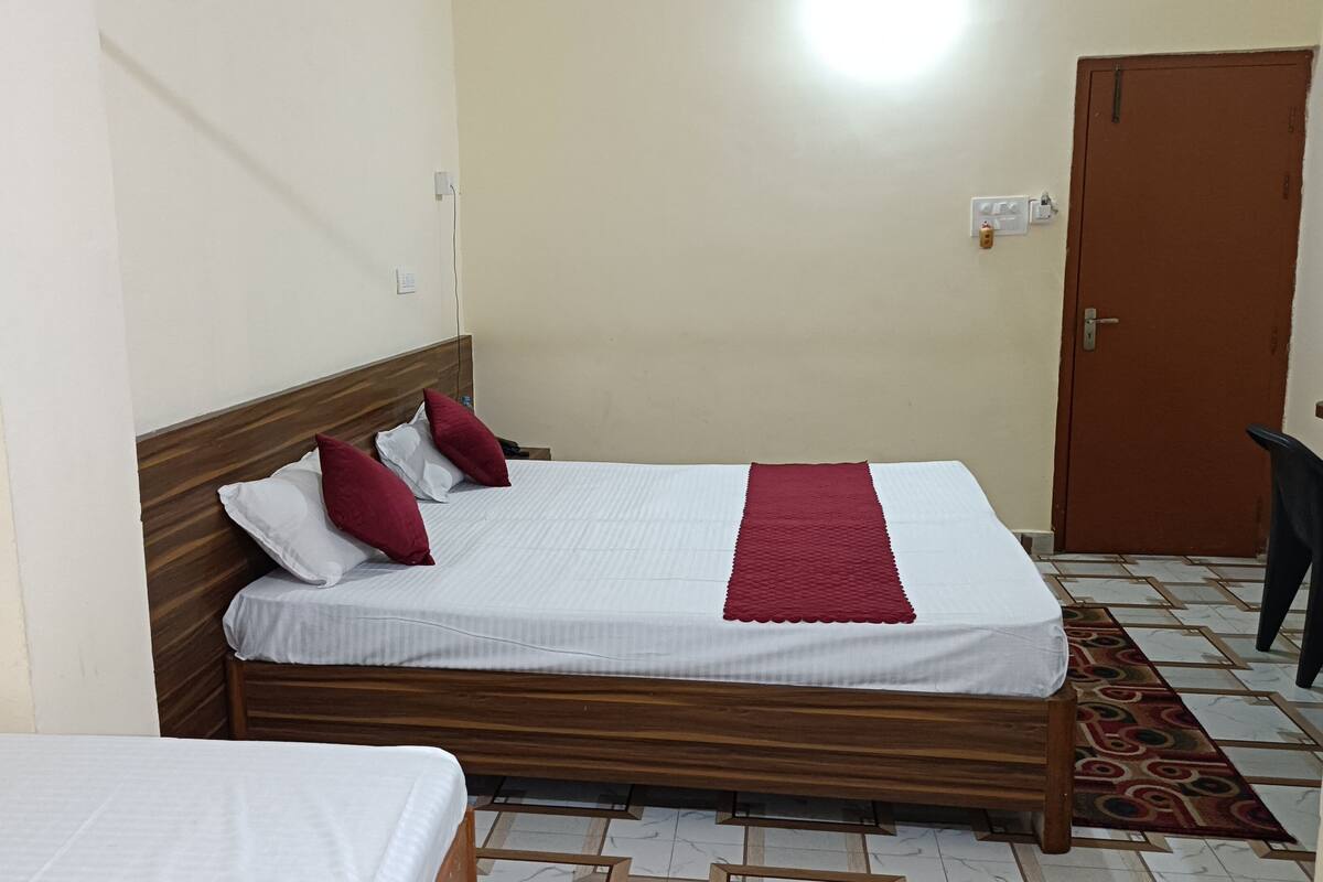 A single bed with a wooden headboard, white bedding, and red accents. A door and a chair are visible in the background.