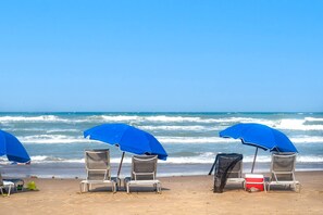 Ein Strand mit vier Metall-Strandstühlen und blauen Sonnenschirmen, wobei einer mit einem schwarzen Tuch bedeckt ist.