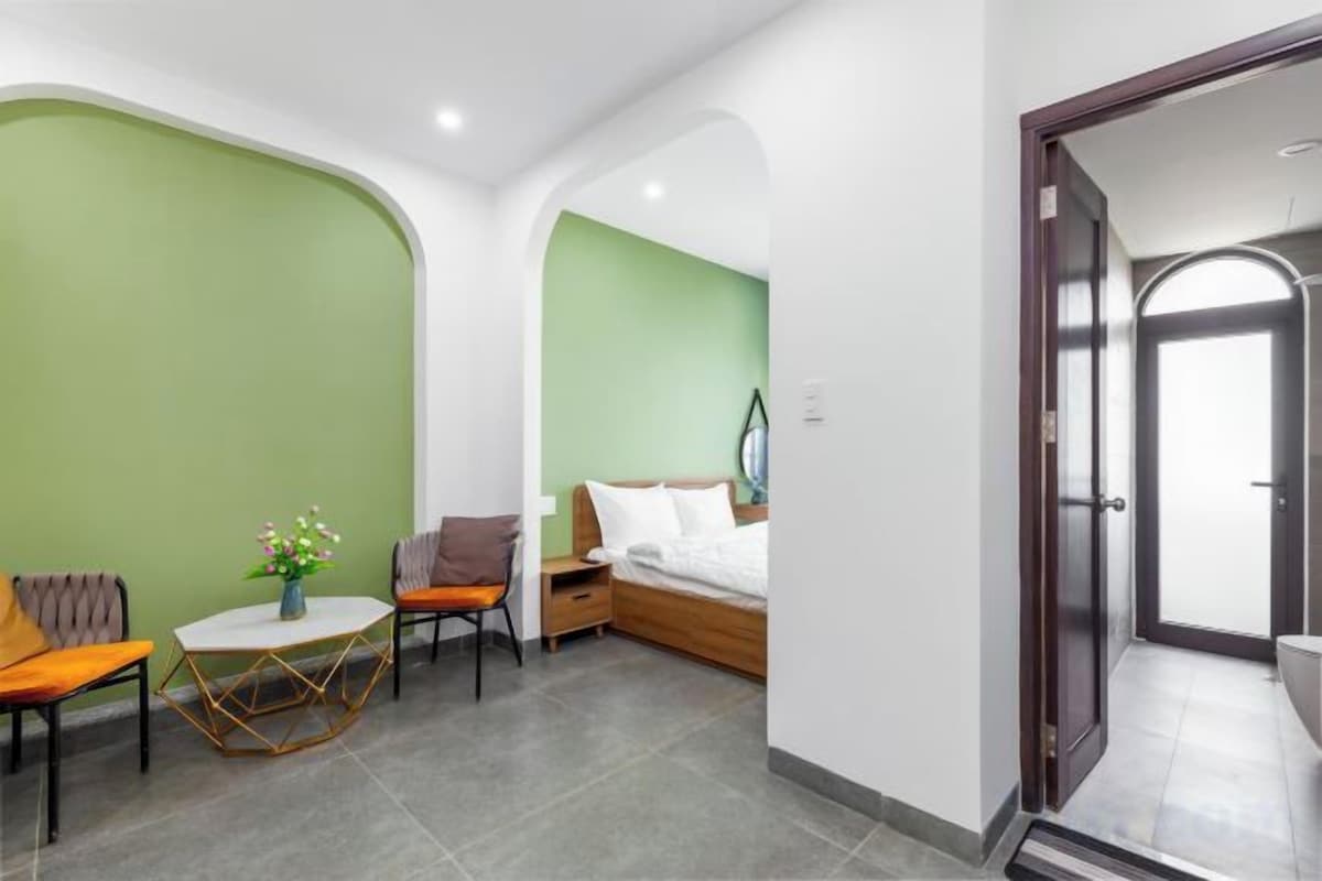 A modern hotel room with a green accent wall, a bed with white linens, a small round table with a vase, and a seating area with two chairs.