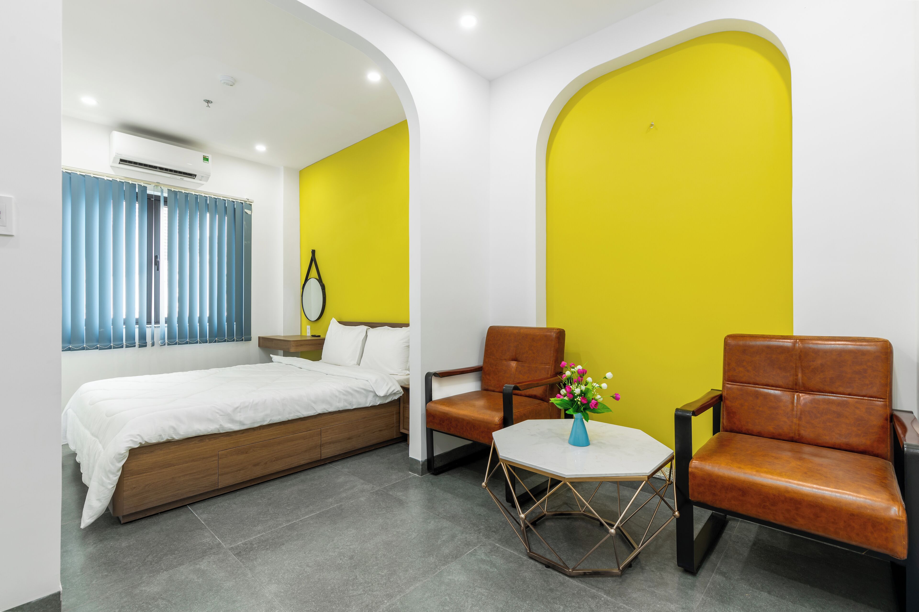 Une chambre d’hôtel moderne avec un lit, deux fauteuils en cuir, une petite table avec un vase et un mur d’accent jaune vif.