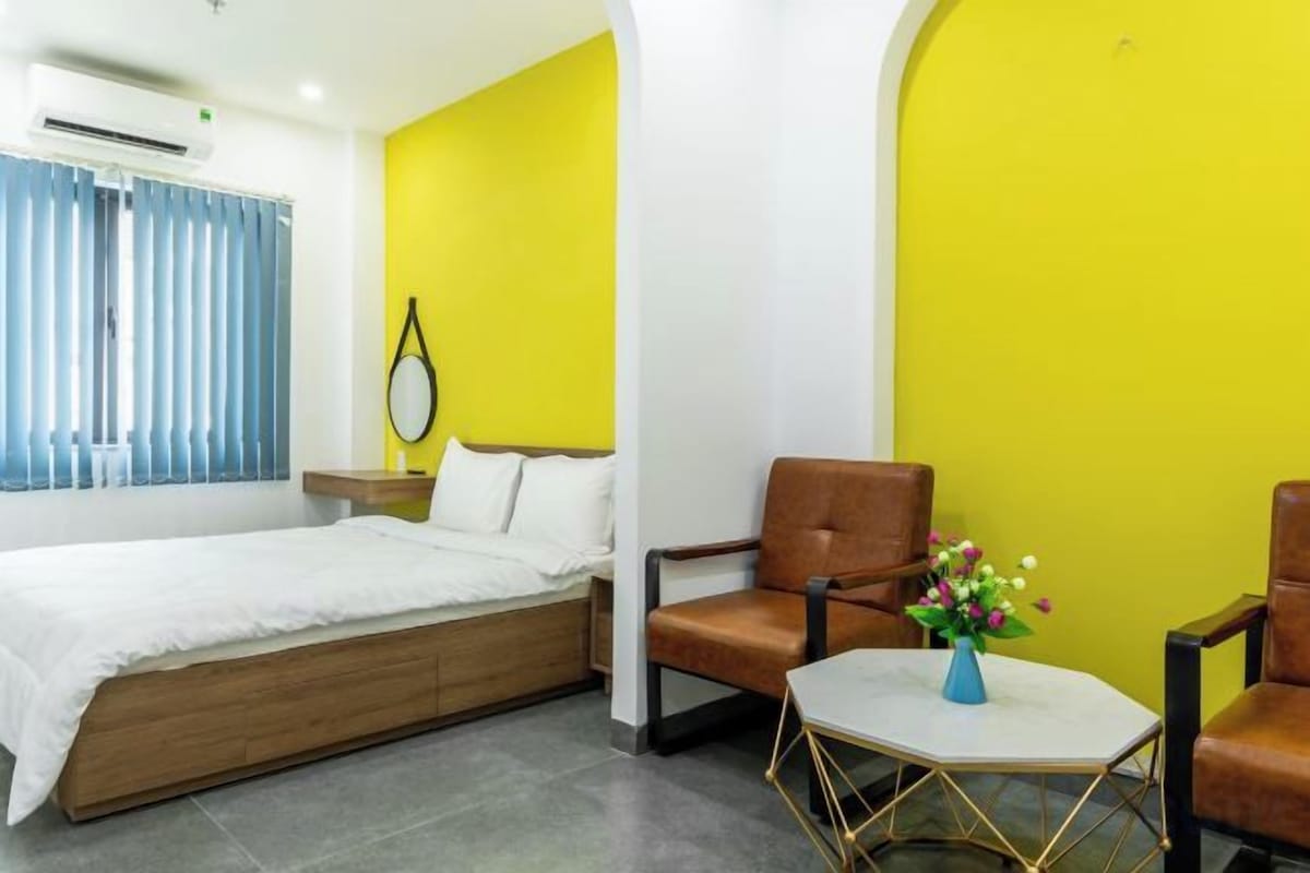 A hotel room with a bed, two chairs, a small table with flowers, and a window with blinds.