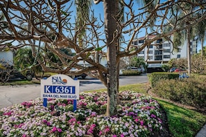 Naamplaatje voor 'ISIA DEL SOL' met adres 'K 6361' en 'BAHIA DEL MAR BLVD', voor een gebouw met geparkeerde auto's en een boom met bloemen.
