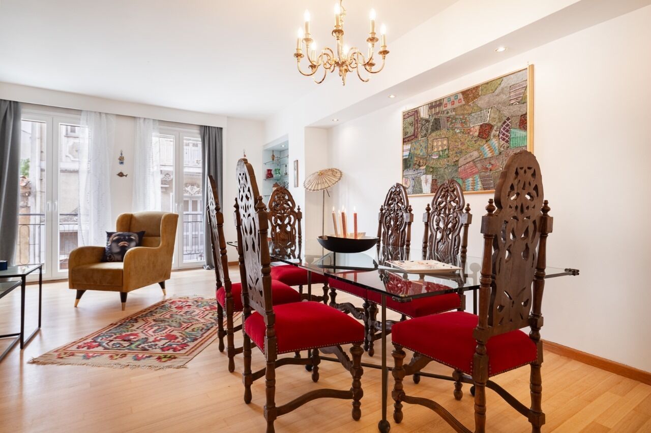 Un comedor con una mesa de vidrio y sillas ornamentadas, una araña y un cuadro abstracto grande en la pared.