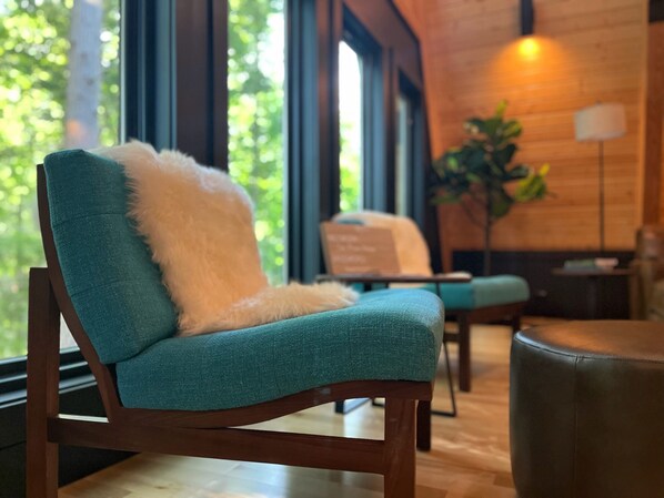 A teal upholstered chair with a white fluffy pillow, positioned in a room with wooden walls and large windows.