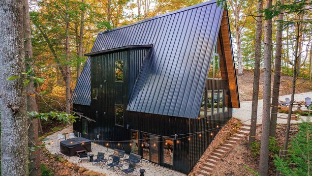 Eine moderne Holzhütte mit geneigtem Dach, großen Fenstern und einer Terrasse mit Sitzgelegenheit im Freien.