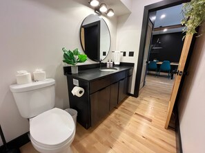 A modern bathroom with a toilet, sink, and mirror. There is a plant on the sink counter and a towel hanging on the wall. - Two Pines Hollow | Private A-Frame | Hot Tub (Rogers)