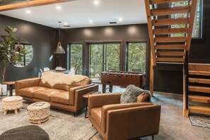 A modern living room with a brown leather sofa, a foosball table, and a wooden staircase.