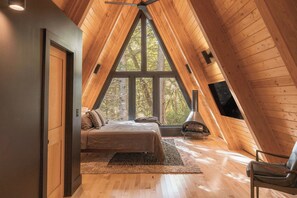 A cozy attic bedroom with a bed, a fireplace, and a large window. - Two Pines Hollow | Private A-Frame | Hot Tub (Rogers)