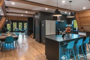 A modern kitchen with a central island, bar stools, and a dining area with blue chairs. - Two Pines Hollow | Private A-Frame | Hot Tub (Rogers)