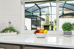 Uma cozinha com bancada branca, uma tigela de frutas, um vaso de planta e uma janela com vista para uma casa.