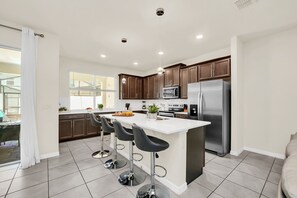 A modern kitchen with a central island, bar stools, stainless steel appliances, and a large window. - 4BR family home - themed rooms & private pool (Kissimmee)