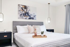 A modern bedroom with a grey headboard, white bedding, a bedside table with a candle, a tray with a bottle and glasses, and a wall-mounted abstract painting. - 4BR family home - themed rooms & private pool (Kissimmee)