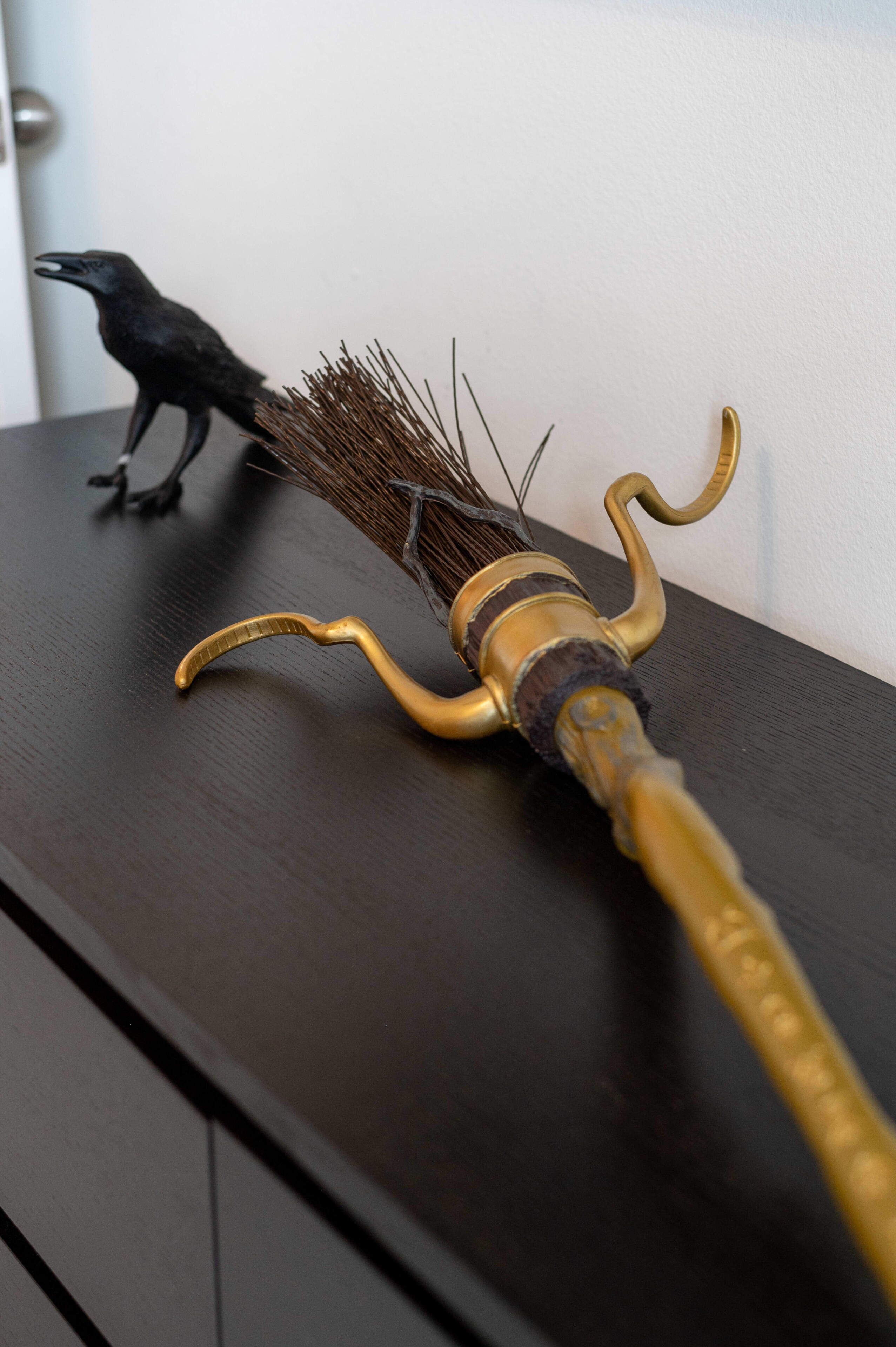 Een bezem met een gouden steel en een zwarte kraakvogel figuur op een donker houten oppervlak.