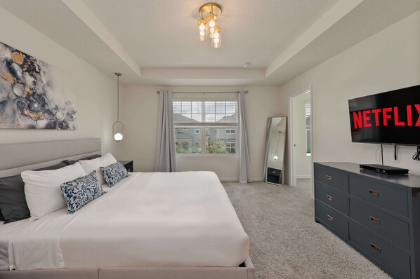 A bedroom with a bed, a TV, a dresser, a window with curtains, and a painting on the wall.