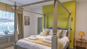 A four-poster bed with white linens, a bedside table with a lamp, and a window with curtains. - The Bat and Ball (Canterbury)