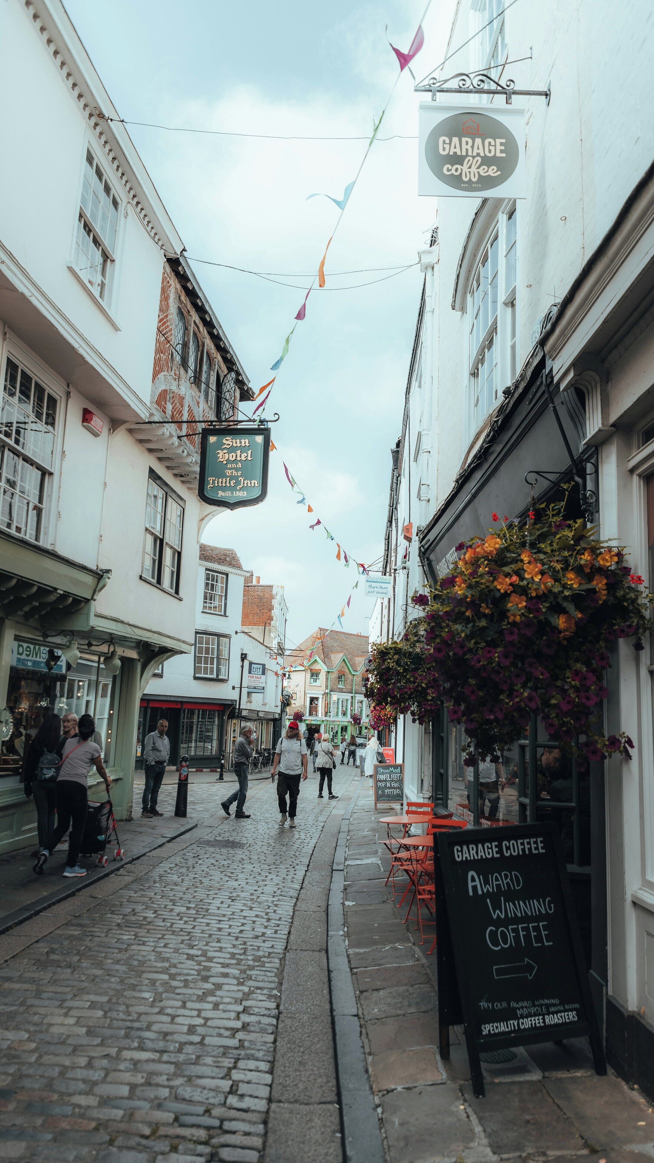 Una calle empedrada con edificios, un cartel de Garage Coffee y un arreglo floral.