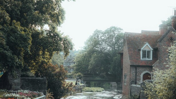 A riverside scene with a brick building, a stone bridge, and a flower-lined path. - The Bat and Ball (Canterbury)