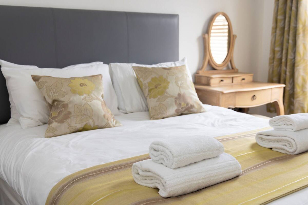 A neatly made bed with white linens, two floral pillows, and two folded towels on a yellow bedspread.