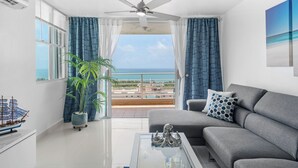 A modern living room with a grey sofa, a coffee table, and a ceiling fan. A large window offers a view of the sea and a balcony. - Perla del Mar | Perfect Beach Escape (Maunabo)