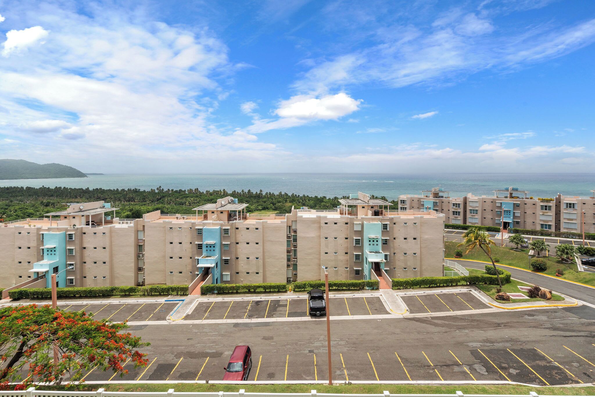 A multi-story hotel with a parking lot in front, a view of the sea, and a hilly landscape in the background.