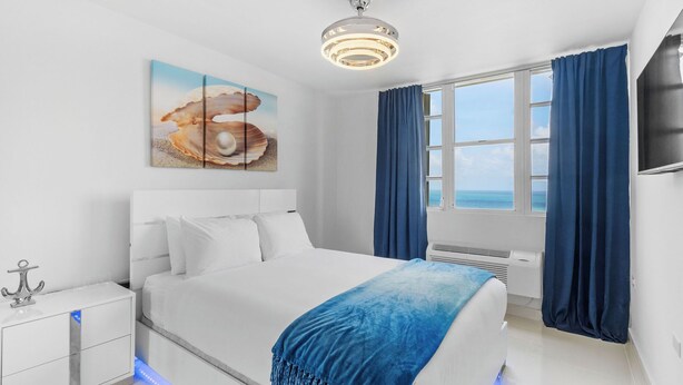 A modern hotel room with a large bed, a nightstand, a ceiling light, a painting, a window with curtains, and a view of the sea. - Perla del Mar | Perfect Beach Escape (Maunabo)