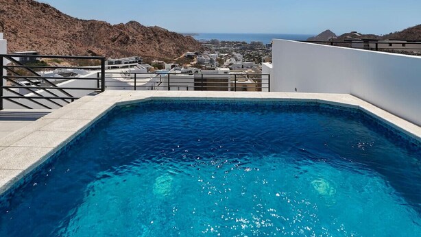 Une piscine à l’eau d’un bleu cristallin, entourée d’une terrasse blanche et d’une rambarde en métal. Au fond, on aperçoit des bâtiments et un paysage montagneux.