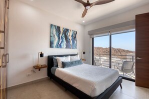Relax in this serene bedroom with a large bed, bedside tables, and a ceiling fan, offering a view of the surrounding landscape from the adjacent balcony.