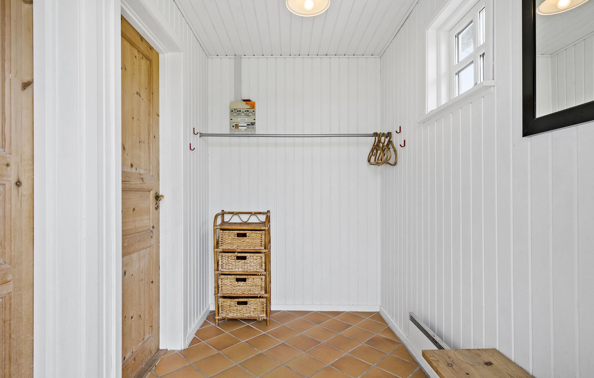 A narrow hallway with white walls, a wooden door, and a tiled floor. There is a metal shelf with a box and a wicker basket.