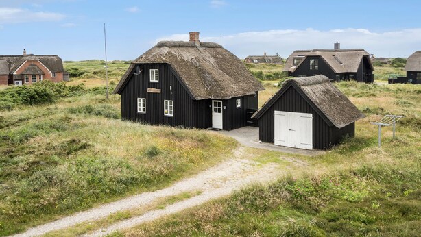 Ein traditionelles, schwarzes Holzhaus mit einem Reetdach, ein kleiner angebauter Schuppen und ein Kiesweg, der zum Eingang führt.