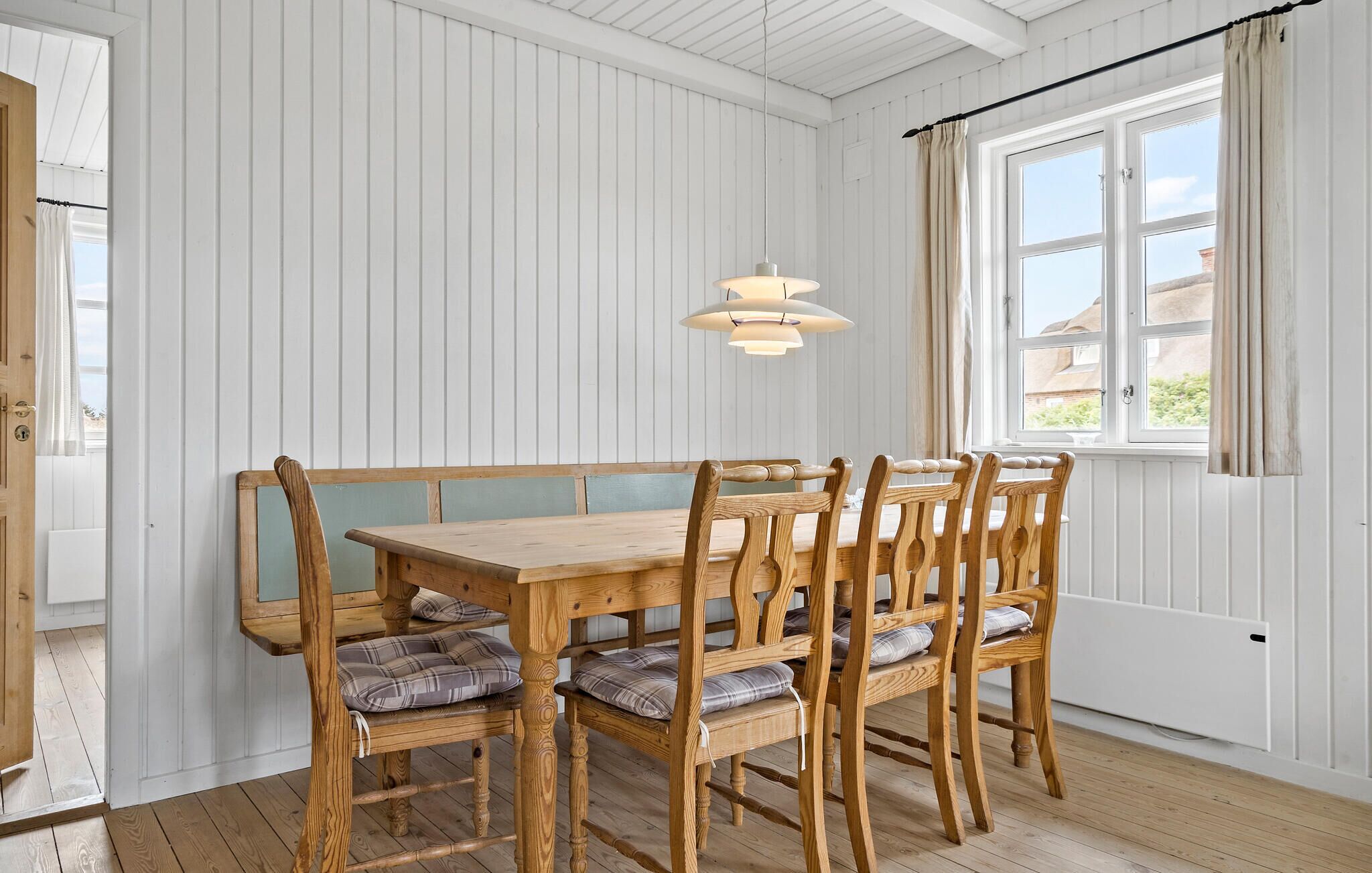 Un coin repas avec une table et des chaises en bois, des murs recouverts de panneaux blancs et une fenêtre avec des rideaux.