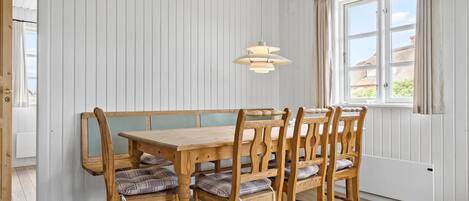Un coin repas avec une table et des chaises en bois, des murs recouverts de panneaux blancs et une fenêtre avec des rideaux.