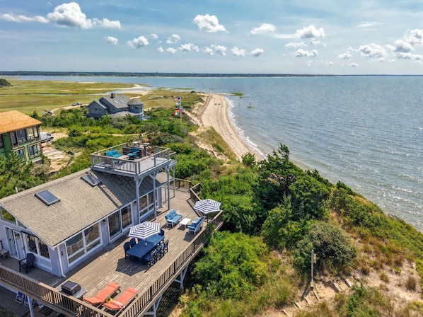 A coastal property with a deck overlooking the beach, a house with a large deck, and a flag. - Hot Tub, Waterfront Home with Stunning Views! (Wellfleet)