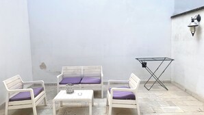 Een klein, afgesloten terras met een witte muur, een raam en een metalen klaptafel met een paars kussen.