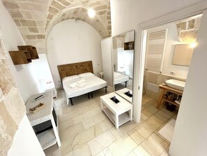 A modern hotel room with a stone arch ceiling, a wooden bed, a white bedside table, a small bedside table with a mirror, a white sink, and a wooden cabinet. - Salto Nel Blu (Leverano)