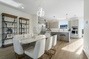 Una cocina moderna con una isla central, armarios blancos y un comedor con mesa de vidrio y sillas blancas.
